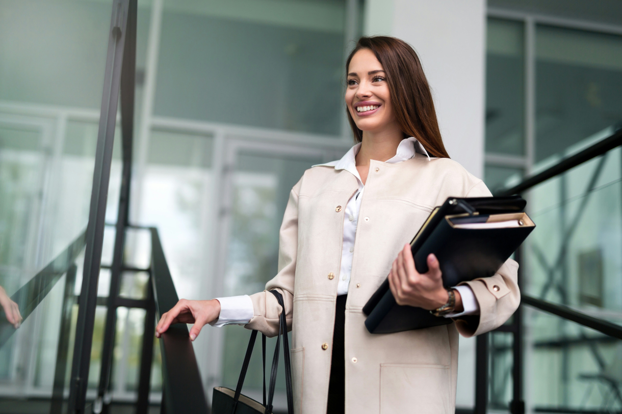 Une femme souriante monte un escalier dans un environnement professionnel moderne. Elle tient des dossiers et un ordinateur portable sous le bras, ainsi qu’un sac à main, montrant une attitude assurée et professionnelle.